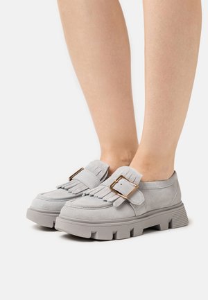 Mocassins en daim gris avec une semelle épaisse, présentant des accents de franges et une boucle décorative. Texture lisse et forme arrondie.