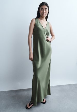 Γυναίκα με μακριά σκούρα μαλλιά, φορώντας αμάνικο μάξι φόρεμα σε ελιά πράσινο χρώμα και μαύρα σαγιονάρες, στέκεται μπροστά σε απλό ανοιχτό φόντο.