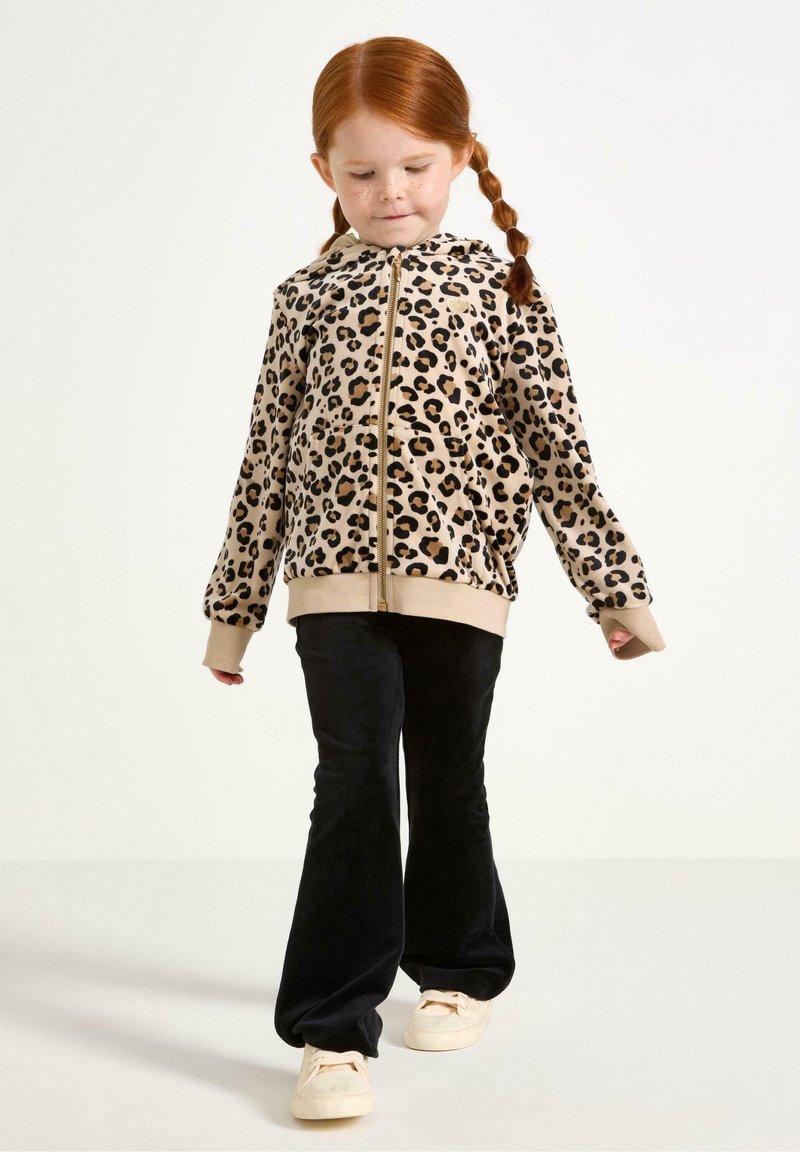 Haut à capuche zippé imprimé léopard en tan avec des taches noires, doté d'un ourlet et de poignets beiges, associé à un pantalon noir évasé et des chaussures blanches.