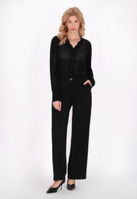 Cardigan nero lavorato a maglia con chiusura a bottoni e design a trama aperta, abbinato a pantaloni neri a gamba larga e tacchi neri.