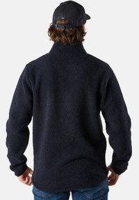 Pullover in pile navy con colletto alto, tessuto testurizzato, maniche lunghe e orlo arrotondato; indossato con un cappellino nero e jeans.