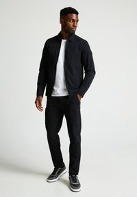 Melnā vieglā jaka ar rāvējslēdzēju priekšpusē, ar divām krūšu kabatām, apvienota ar melnām biksēm, kam ir rāvējslēdzēja sānu kabatas un pelēkiem sporta apaviem.