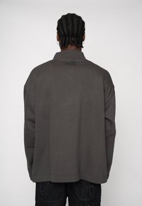 Nadmierny, ciemnoszary sweter z kołnierzem, subtelną fakturą tkaniny i logo na plecach. Gładka powierzchnia i luźny krój.