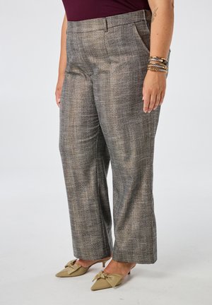 Personne portant un pantalon large à carreaux gris, des talons pointus beiges avec des nœuds, et des bracelets en or au poignet droit, debout devant un fond blanc.