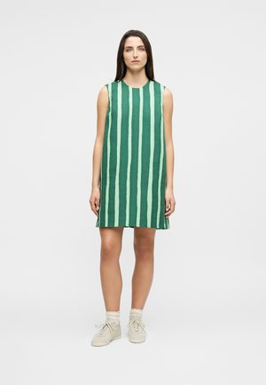 Žena stojaca v zelených a svetlozelených vertikálne pruhovaných šatách bez rukávov, obutá v bielych ponožkách a béžových teniskách, na jednoduchom pozadí.