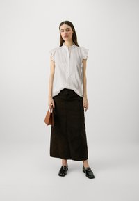 Blusa blanca de manga corta con acentos de volantes, combinada con una falda larga de mezclilla marrón oscuro. La modelo sostiene un bolso marrón. Se ven unos mocasines negros.