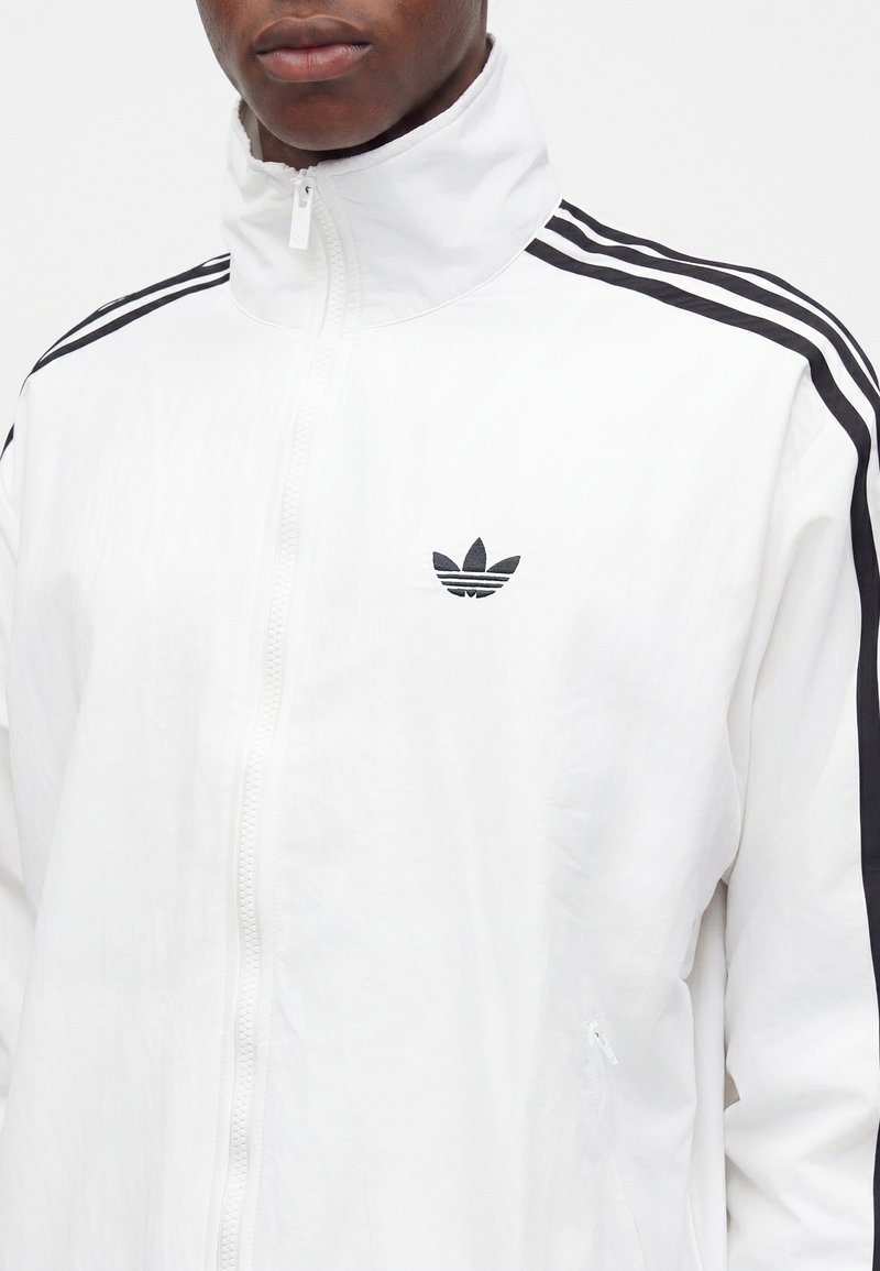 Mand iført en hvid lynlåstrøje med sort Adidas-logo på brystet og tre sorte striber ned ad ærmerne.