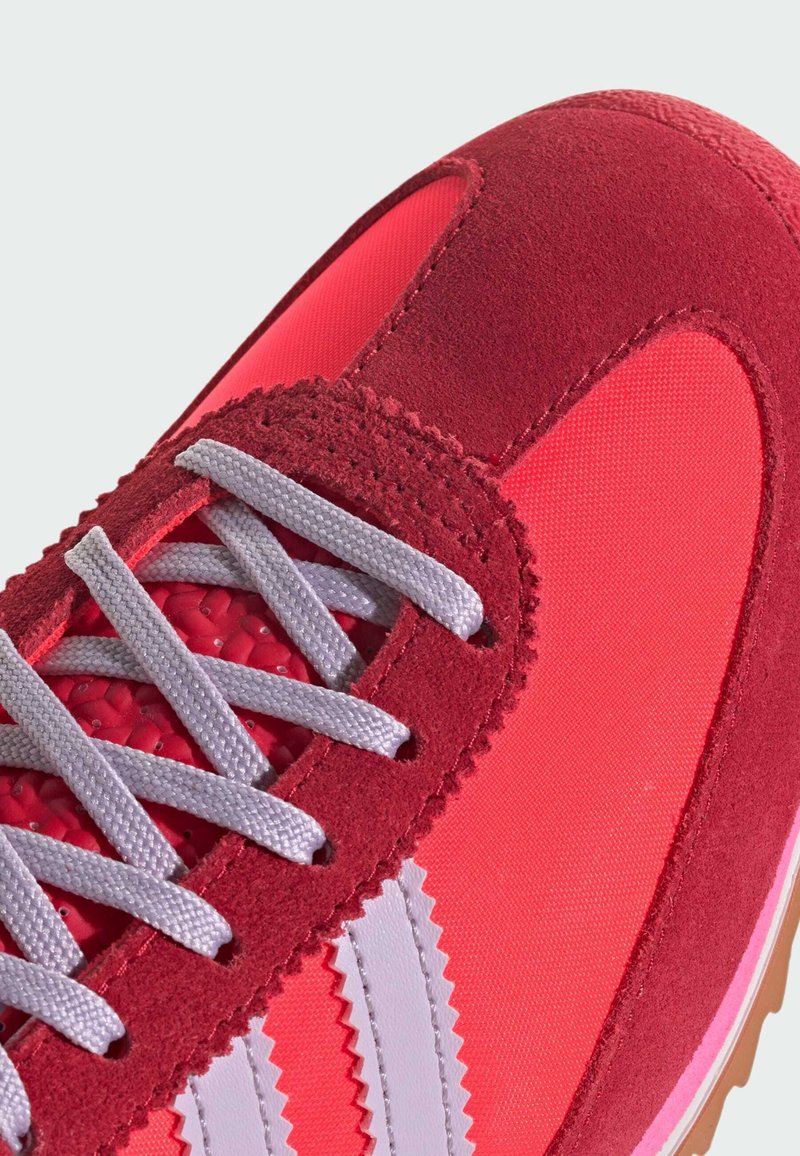 Zapatilla deportiva roja y rosa con parte superior de ante y malla, que presenta cordones grises y rayas serradas blancas en los laterales. Acentos texturizados visibles.