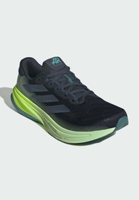 Schwarzer Sportschuh mit Mesh-Obermaterial, grünen Akzenten und drei Streifen-Logo. Verfügt über eine abgerundete Zehenpartie und eine gepolsterte Sohle für Unterstützung.