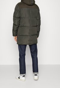 Padded dark green parka with a hood, featuring a contrasting brown panel on the shoulders and a quilted design. Worn over blue jeans and white sneakers.