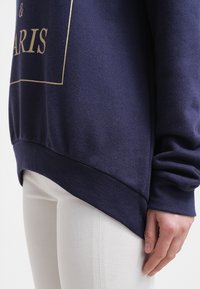 Felpa blu navy con lettering dorato, dotata di maniche lunghe, vestibilità rilassata e orlo curvo, abbinata a pantaloni bianchi.