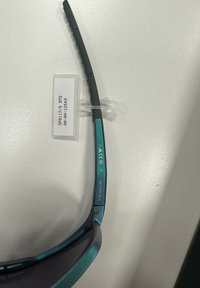 Sluneční brýle s tyrkysovým kovovým rámečkem a šedými stranami. Čočky s přechodem barvy od tmavé po světlou. Označení obsahuje "NENÍ NA PRODEJ", CE a detaily modelu.