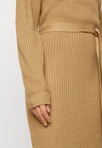 Gros plan d'une personne portant une robe-pull en tricot beige avec une ceinture assortie nouée à la taille et des manches longues couvrant le poignet.