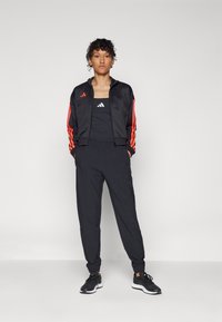 Zwarte trainingspak met een ritsjack met rode strepen en logo, gecombineerd met een zwart tanktop en bijpassende joggerbroeken. Zwarte sneakers.