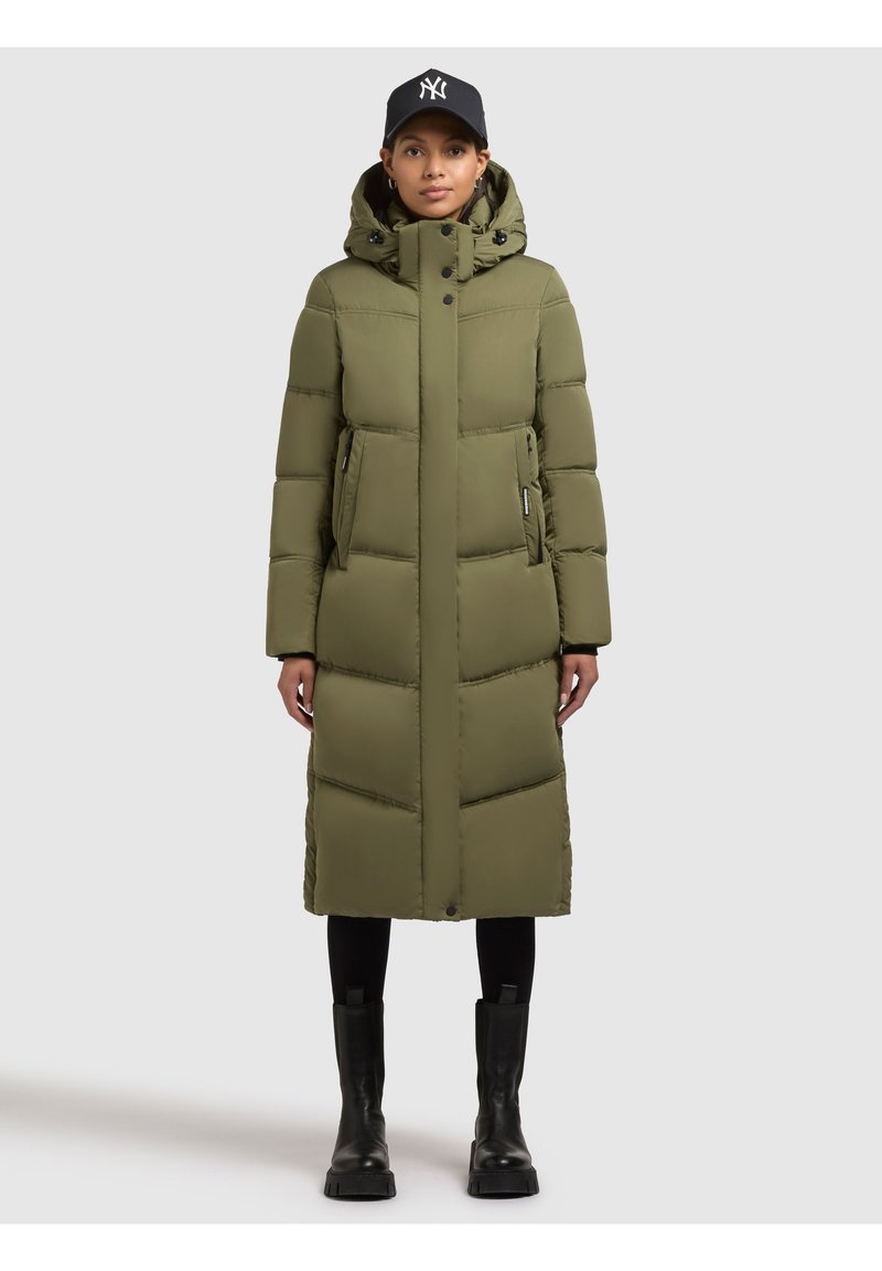 Olivinvihreä toppatakki hupulla, jossa on pitkä siluetti, vetoketjulliset sivutaskut ja resorit hihansuuissa. Yhdistetty mustiin housuihin ja saappaisiin.