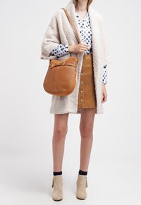 Borsa a spalla in pelle color cuoio con forma arrotondata, dettaglio con fibbia e tracolla regolabile, abbinata a un cappotto bianco morbido e una gonna in camoscio.