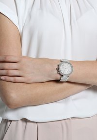 Silver-tone watch with a round face, crystal accents, and a metal bracelet. Positioned on a wrist, with a light-colored blouse in the background.