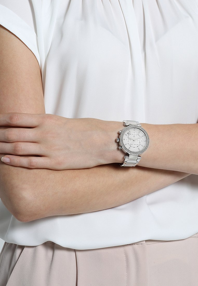 Silver-tone watch with a round face, crystal accents, and a metal bracelet. Positioned on a wrist, with a light-colored blouse in the background.