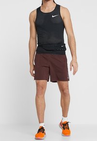 Hombre que lleva una camiseta sin mangas negra de malla, pantalones cortos burdeos, una pouch negra en la cintura, calcetines blancos y zapatillas de correr naranjas brillantes con cordones negros.