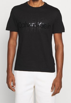 Mężczyzna w czarnym t-shircie z krótkim rękawem z nałożonym logo Calvin Klein i białych spodniach, na jednolitym tle.