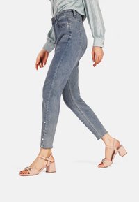 Jeans azzurri chiari a vita slim, con bottoni decorativi sulle caviglie. Abbinati a sandali con tacco a blocco rosa pallido.