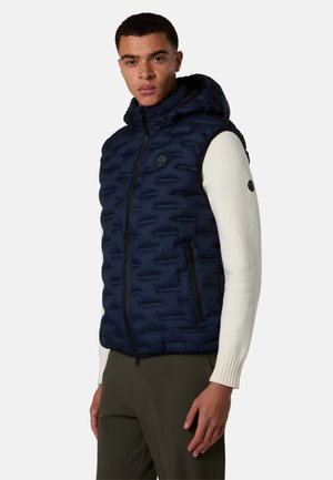 Mand iført en navy polstret hættetrøje over en hvid langærmet sweater og olivengrønne bukser, stående mod en ensfarvet baggrund.