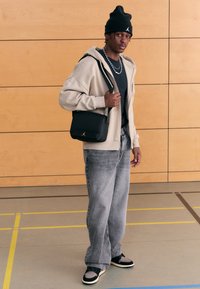 Beige hettejakke med glidelås over en svart t-skjorte, grå jeans, svart lue og en svart skulderveske med en gyllen logo. Står på gulvet i et treningsstudio.