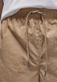Beige shorts med elastisk midja, som dras på, med snörning och silverfärgade metalldetaljer. Tyget har en skrynklig textur.