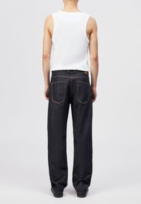 Personne portant un débardeur blanc et un jean denim bleu foncé ample, debout de dos sur un fond clair uni.