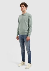 Polo in lichtgroene katoen, met een halflange rits, lange mouwen en geribbelde manchetten. Gecombineerd met strakke blauwe jeans en crèmekleurige sneakers.