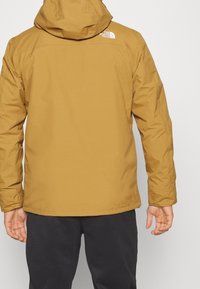 Person i en senapsgul huva-jacka med The North Face-logotyp på övre delen av ryggen, i kombination med svarta byxor, står med ryggen mot kameran.