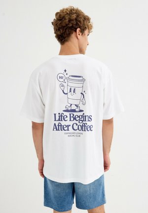 Joven con camiseta blanca que lleva un gráfico de una taza de café caminando y el texto "La vida comienza después del café, Club social de amantes del espresso" en la espalda.