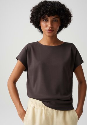 Vrouw met krullend haar die een donkerbruin shirt met korte mouwen en lichtbeige broek draagt, staand met handen in de zakken tegen een effen achtergrond.