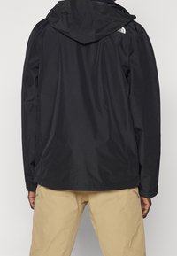 Černá voděodolná bunda s kapucí, která má uvolněný střih, jednoduchý design a logo na rameni. V kombinaci s béžovými kalhotami.