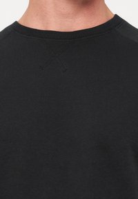 Gros plan sur le cou et le haut de la poitrine d'une personne portant un sweat-shirt noir uni à col rond avec un détail "X" cousu sous le col.