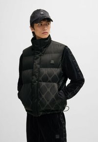Black puffer vest with high collar, featuring quilted pattern and logo. Accented with plush black sleeves and a subtle repeating design.
