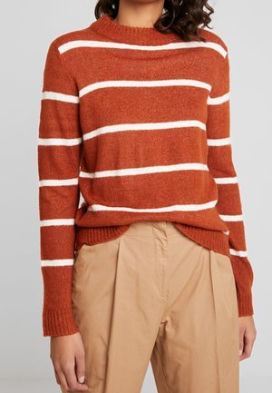 Mujer con suéter de punto color óxido con rayas horizontales blancas y pantalones beige de talle alto y plisados, frente a un fondo gris.
