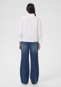 Bluză albă cu un guler înalt, volanat și mâneci bufante, asociată cu pantaloni largi din denim albastru decolorat, moale la atingere.