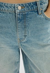 Närbild av ljusblå denimjeans med orange sömmar och en metallknapp graverad med "BERSHKA DENIM."