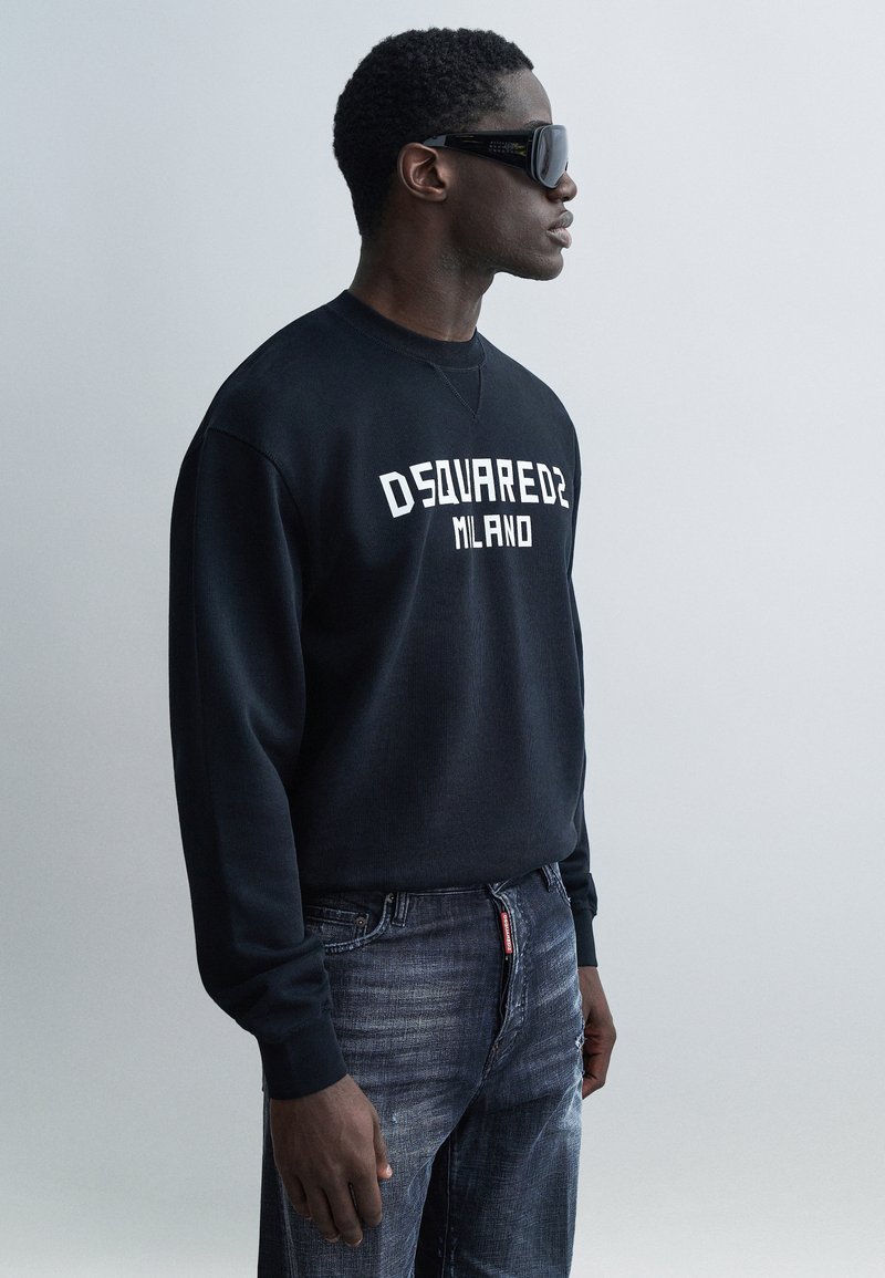 Schwarzer Sweatshirt mit weißem "DSQUARED2 MILANO"-Logo, kombiniert mit grauen, abgenutzten Jeans, präsentiert vor hellem Hintergrund.