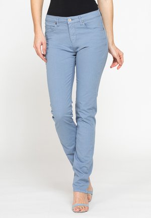 Donna con pantaloni slim fit azzurri chiaro e sandali open toe abbinati, in piedi con le gambe incrociate, indossa un top nero.