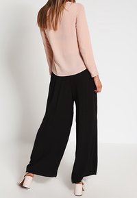 Femme portant un chemisier rose clair à manches longues et un pantalon noir à jambes larges, debout de dos face à la caméra sur un fond blanc.