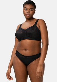 Žena v černé krajkové podprsence s nastavovatelnými ramínky a ladících krajkových kalhotkách, stojící na prostém světle šedém pozadí.