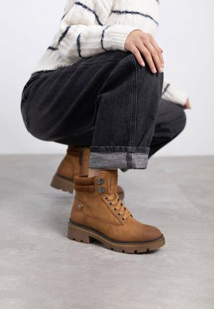 Botas de tobillo marrones de ante con un acabado texturizado, ojales de metal y una suela gruesa de contraste. Combinadas con unos vaqueros grises remangados.