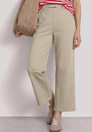 Kvinde iført beige bukser med brede ben, beige sneakers, rød- og hvidstribet top, holder beige ruskind skuldertaske.