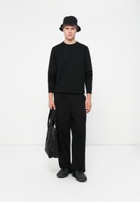 Sort langærmet crewneck sweater parret med sorte cargo bukser, en sort bucket hat og sorte teksturerede sneakers, mens der holdes en sort skuldertaske.