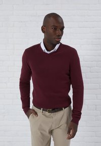Pulover burgund cu decolteu în V, cu manșete înfășurate și design ajustat, purtat cu pantaloni bej și o curea maro, pe un fundal de cărămidă albă.