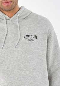 Lys grå hettegenser med snor, som har «NEW YORK» i svart tekst og tre baseballikoner brodert på brystet.