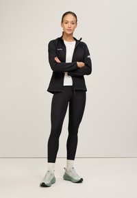 Giacca sportiva nera con cappuccio, chiusura a zip e maniche lunghe sopra leggings neri. Modello che indossa sneakers verde chiaro con dettagli bianchi.