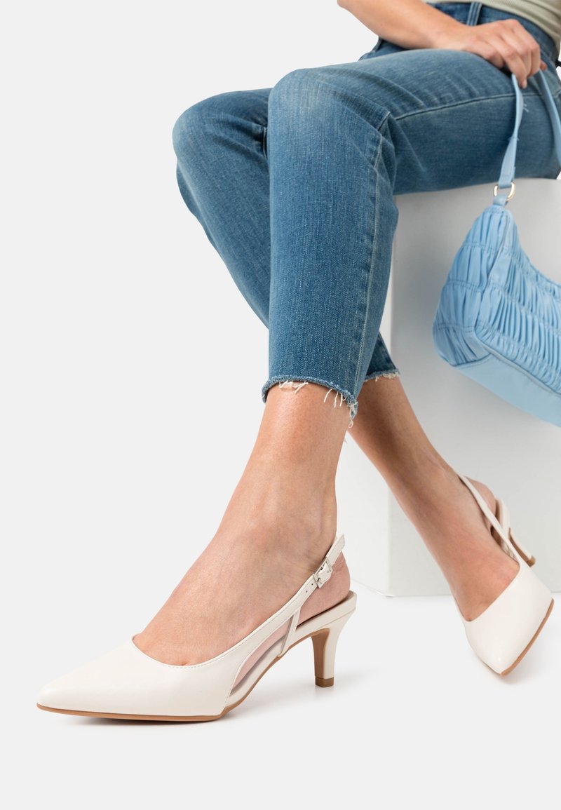 Personne portant un jean skinny bleu clair et des escarpins à bride blanche à talons bas, tenant un petit sac à main bleu clair froncé, assise.